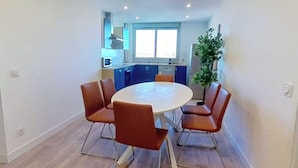 Un coin repas avec une table ronde et six chaises, une cuisine en arrière-plan et une plante en pot.