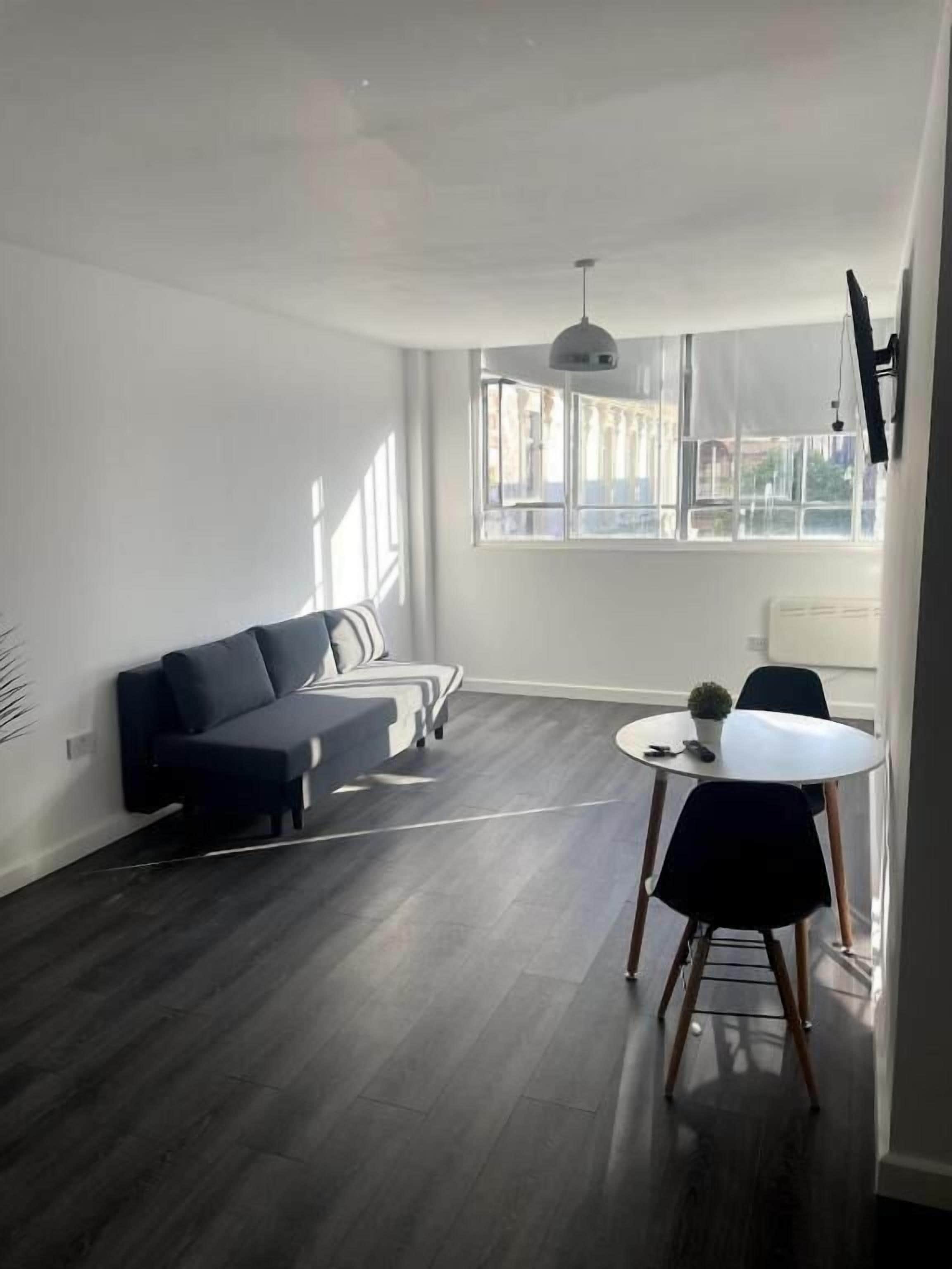 Un salón moderno con piso de madera oscura, un sofá negro, una mesa redonda de comedor con sillas y un gran ventanal que proporciona luz natural.
