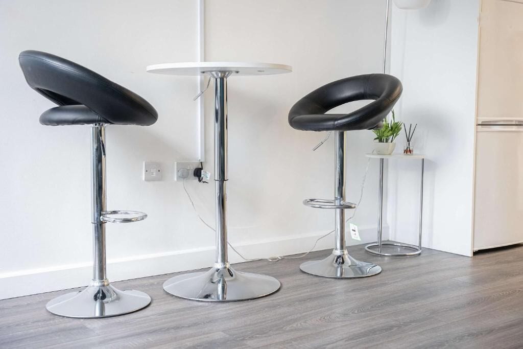 Three modern bar stools with black seats and chrome bases, positioned in a kitchen area with a white wall and a small plant on a shelf.