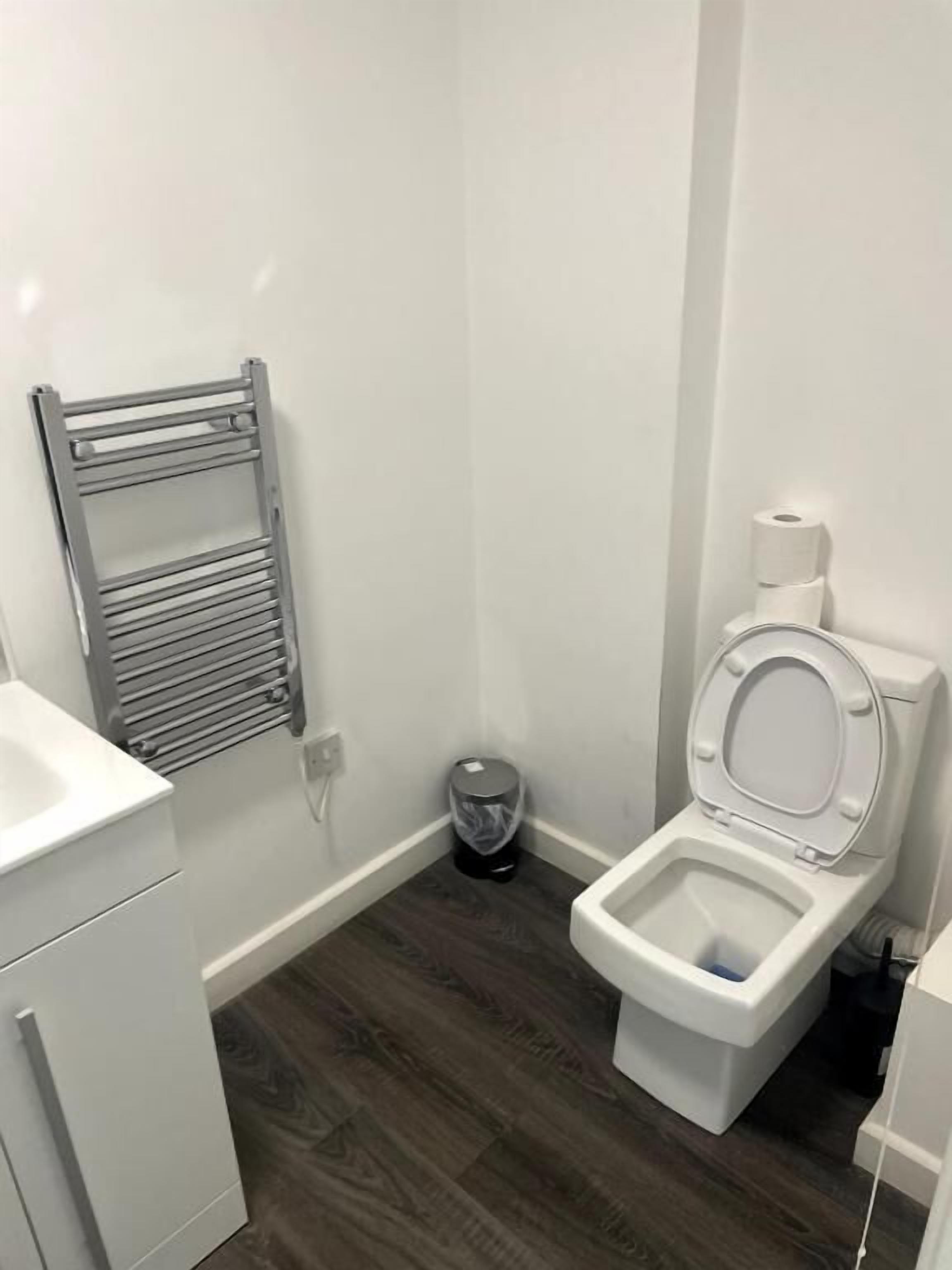 Un baño con inodoro blanco, toallero de metal, lavabo blanco y un cubo de basura.