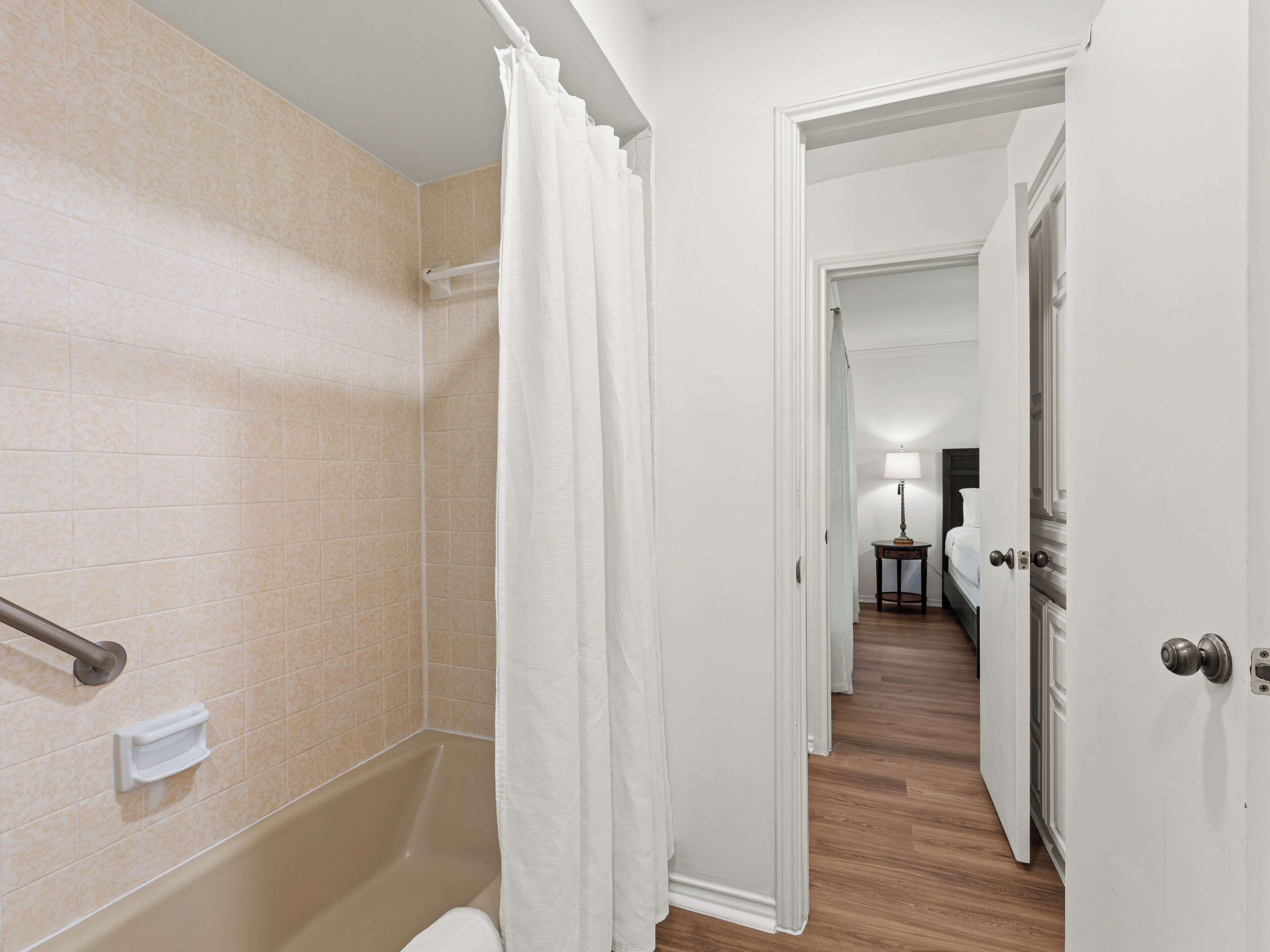 Ein Badezimmer mit sandfarben gefliesten Wänden, einem weißen Duschvorhang, einer Haltestange und einem Holzboden, der zum Schlafzimmer mit Nachttisch und Lampe führt.
