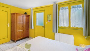 Une pièce aux murs jaunes, avec une armoire en bois, un lit recouvert d’une couvre-lit à motifs floraux, une fenêtre avec des rideaux et un radiateur.