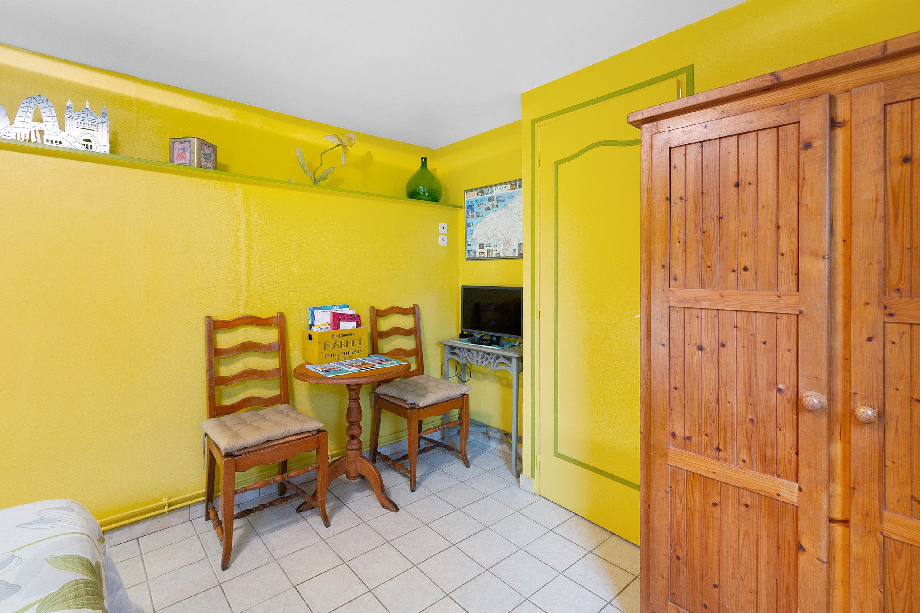 Una habitación pequeña con paredes amarillas, dos sillas de madera, una mesita, un armario de madera y un televisor montado en la pared.