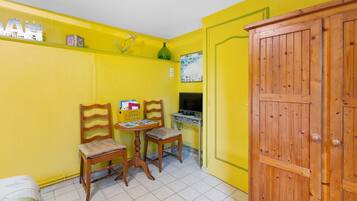Una habitación pequeña con paredes amarillas, dos sillas de madera, una mesita, un armario de madera y un televisor montado en la pared.