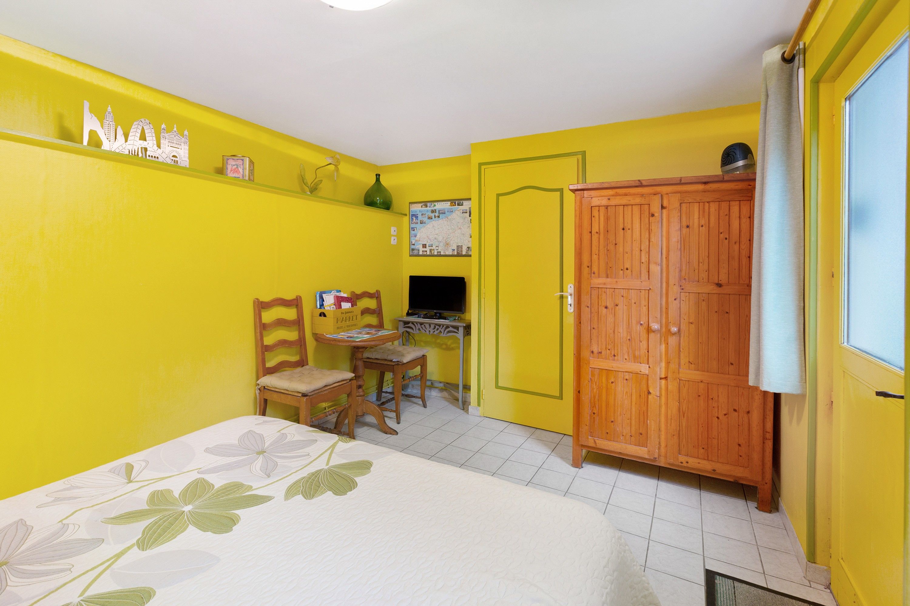 Une pièce aux murs jaunes, avec une armoire en bois, un lit recouvert d’une courtepointe à motifs floraux, une chaise en bois, une petite table et une carte accrochée au mur.