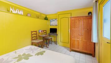 Une pièce aux murs jaunes, avec une armoire en bois, un lit recouvert d’une courtepointe à motifs floraux, une chaise en bois, une petite table et une carte accrochée au mur.