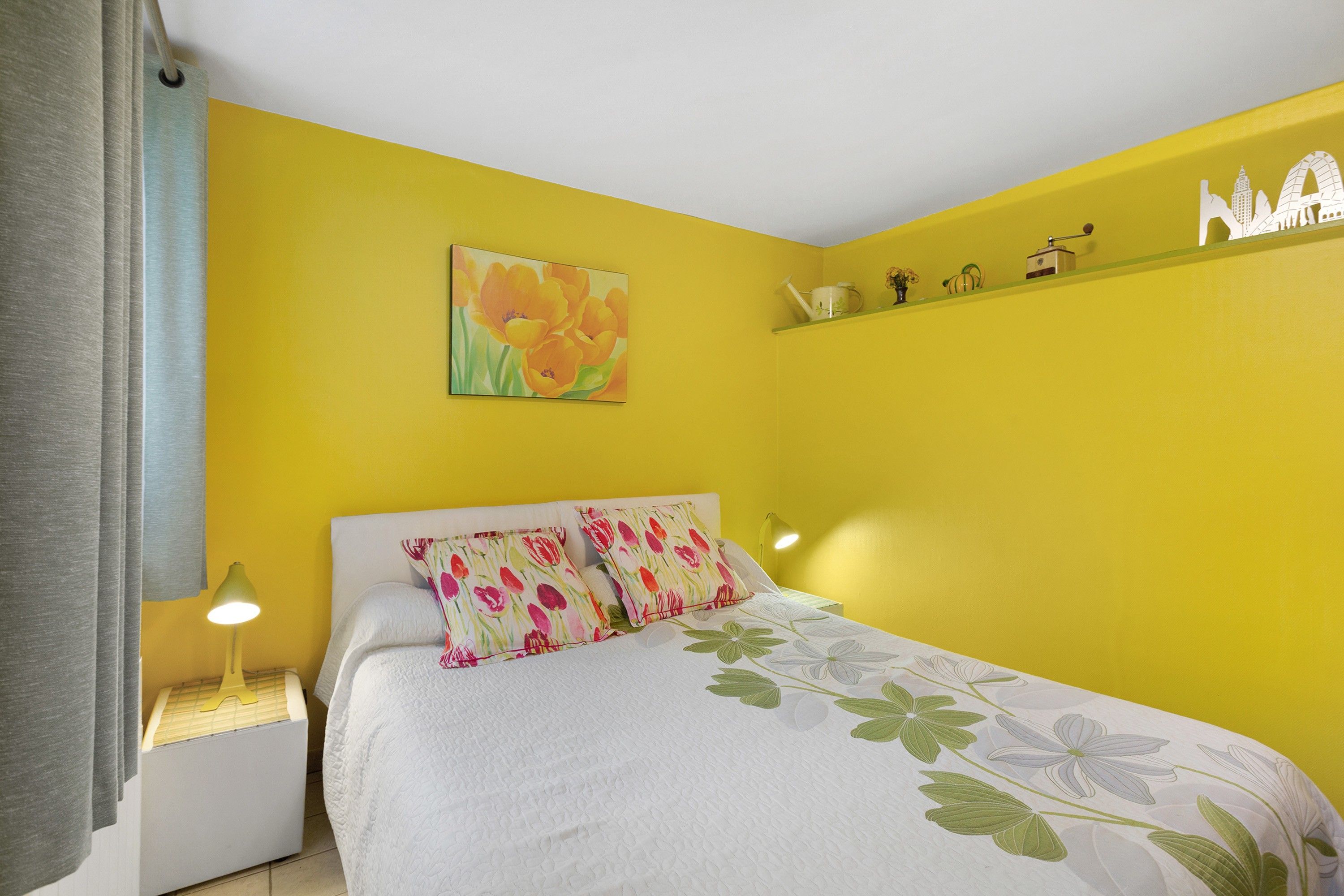 Een slaapkamer met een gele wand, een bed met bloemenbeddengoed, een schilderij van bloemen en een plank met kleine objecten.