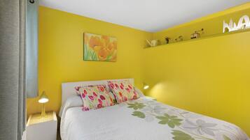 Een slaapkamer met een gele wand, een bed met bloemenbeddengoed, een schilderij van bloemen en een plank met kleine objecten.