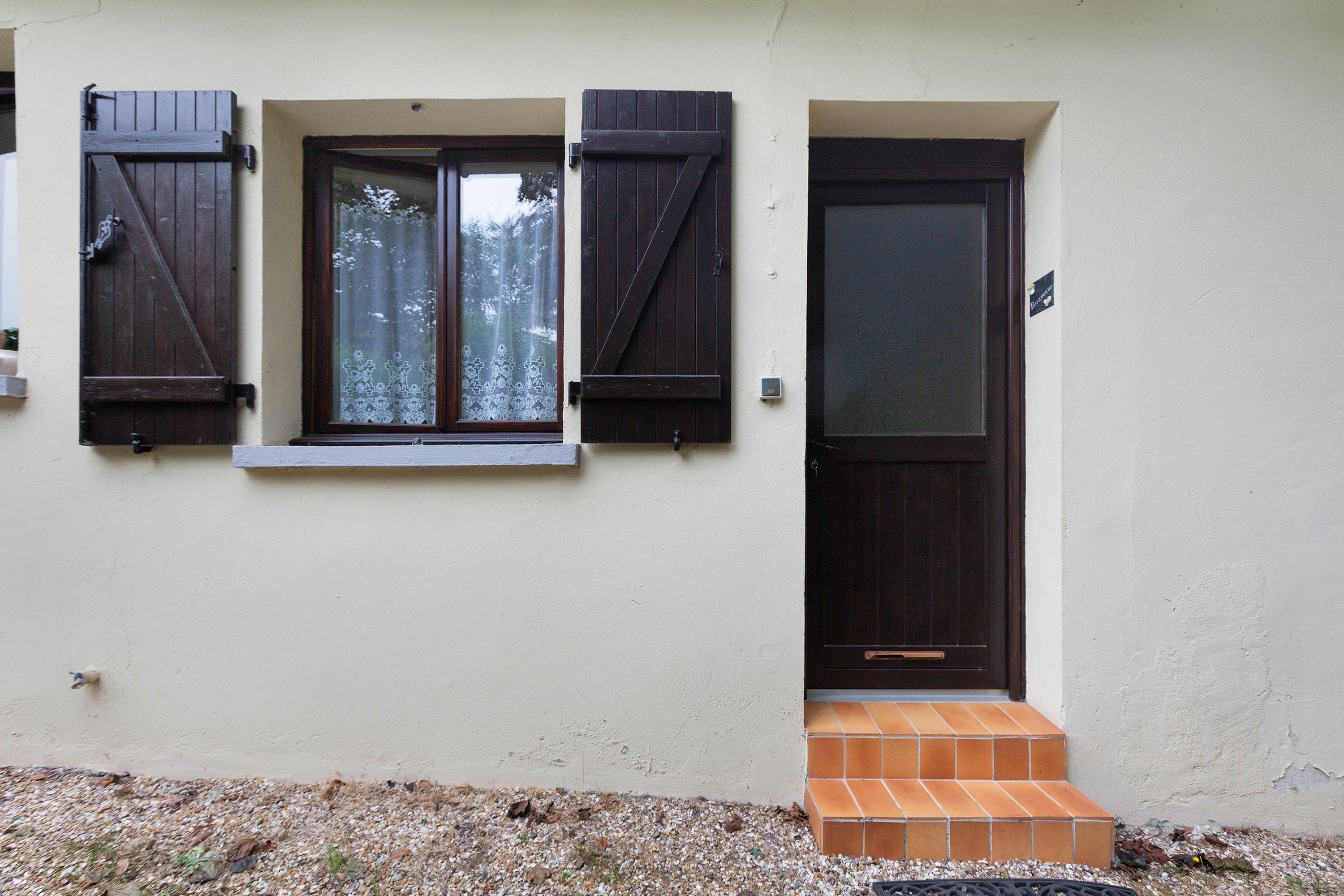 Ein Gebäude mit einer dunklen Holztür, einem Fenster mit Fensterläden und einer kleinen Treppe zum Eingang.