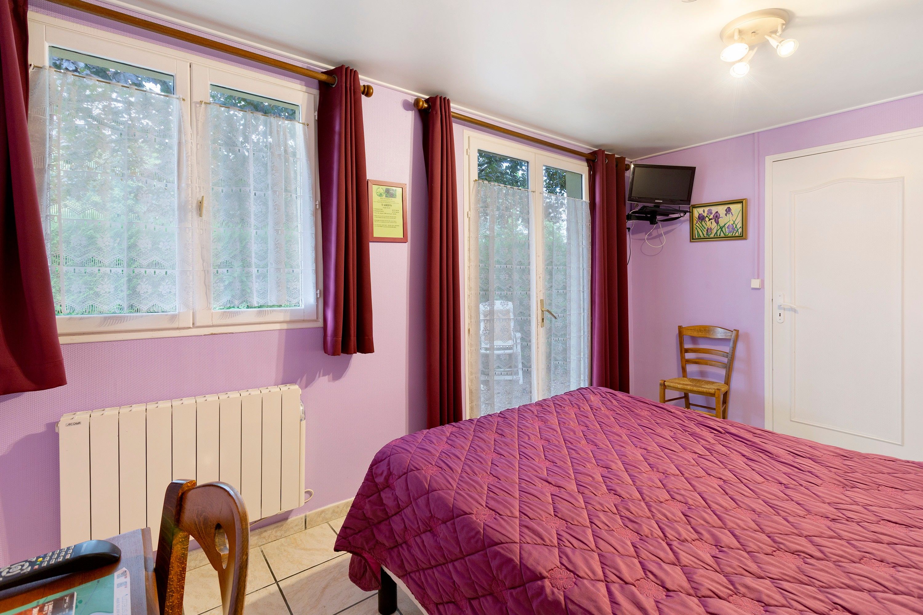 A bedroom with a bed, a chair, a radiator, a television mounted on the wall, and a window with curtains.
