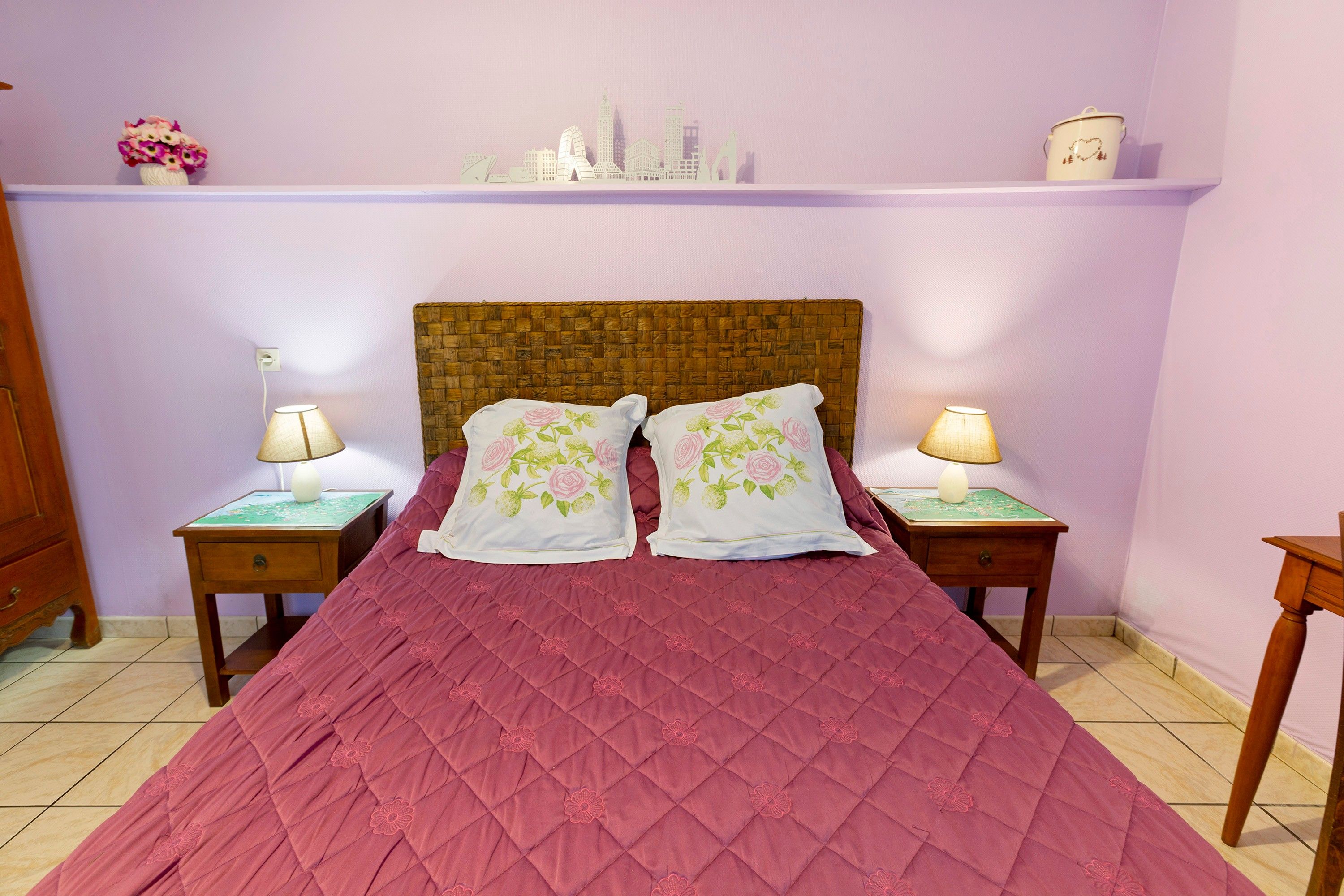Une chambre à coucher avec une tête de lit en bois, une couvre-lit rose matelassée, deux coussins assortis ornés de broderies florales, deux tables de chevet avec des lampes et une étagère avec des objets décoratifs.
