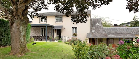 A two-story house with a front lawn, a large tree, and a variety of plants and flowers.