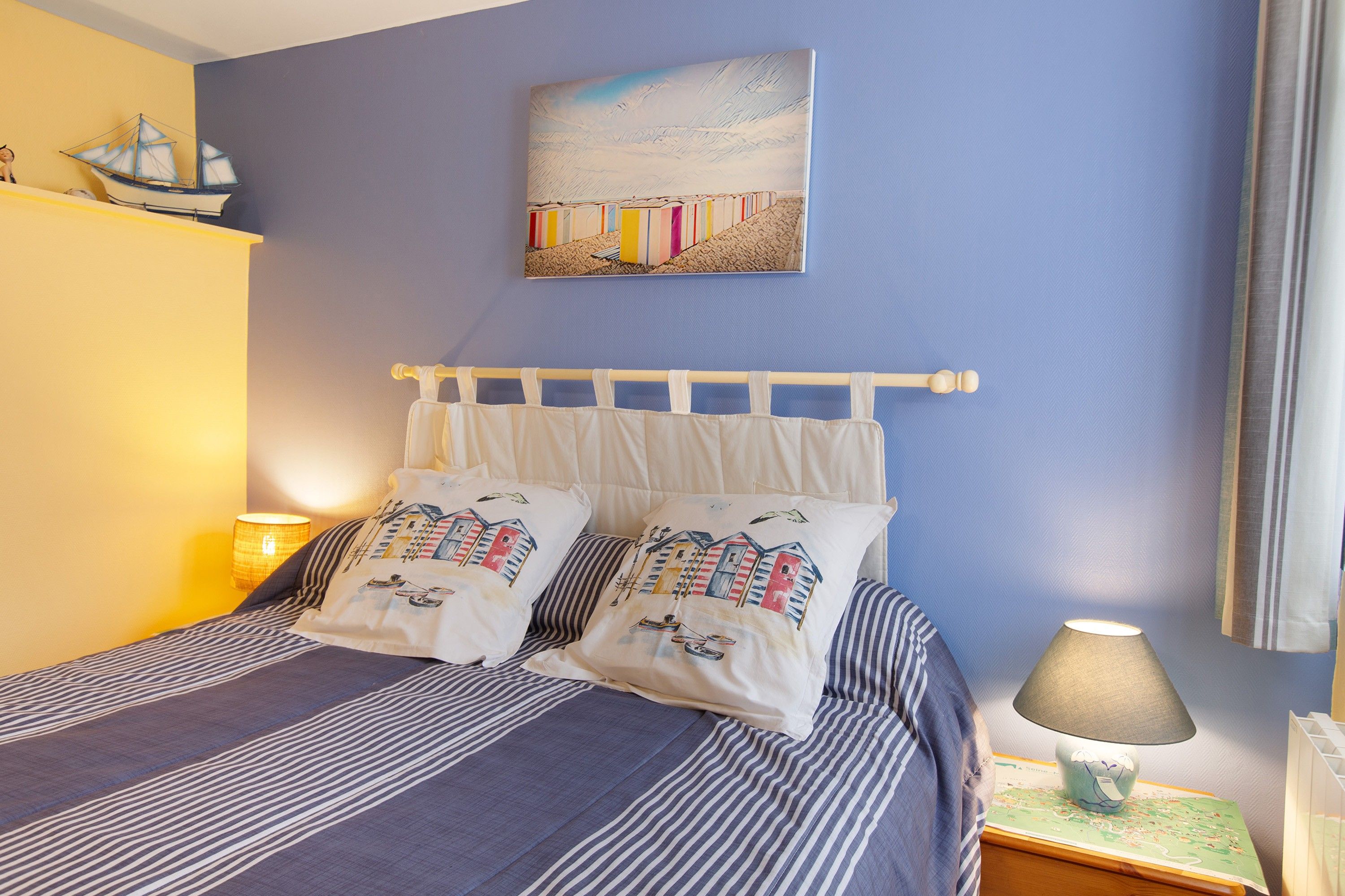 Een slaapkamer met een bed, kussens, een nachtkastje, een lamp, een kaart en een schilderij aan de muur.