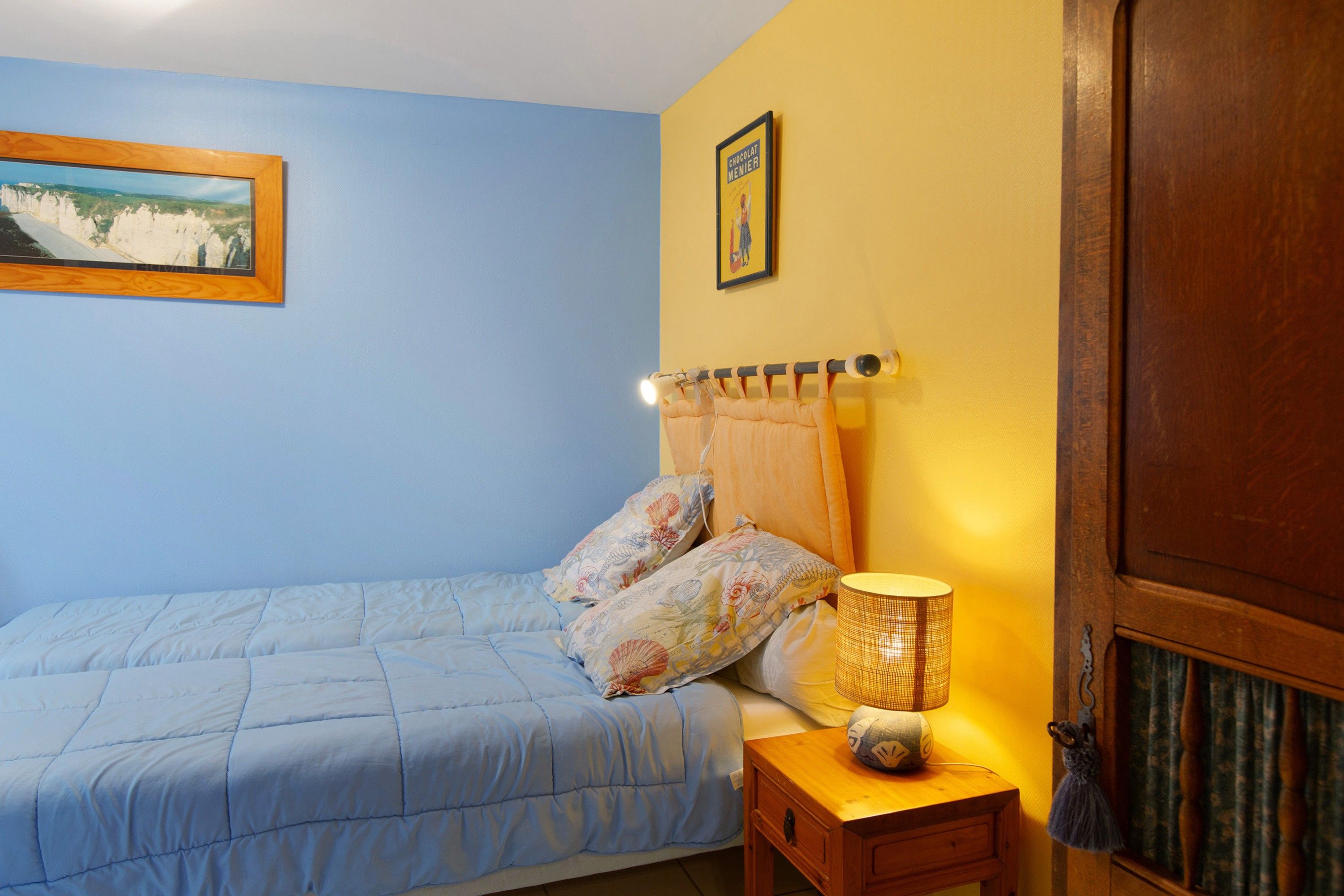 Een slaapkamer met een bed, een nachtkastje, een lamp, een deur en twee afbeeldingen in lijsten aan de muur.