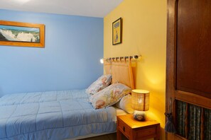 Un dormitorio con cama, mesita de noche, lámpara, una puerta y dos cuadros en la pared.