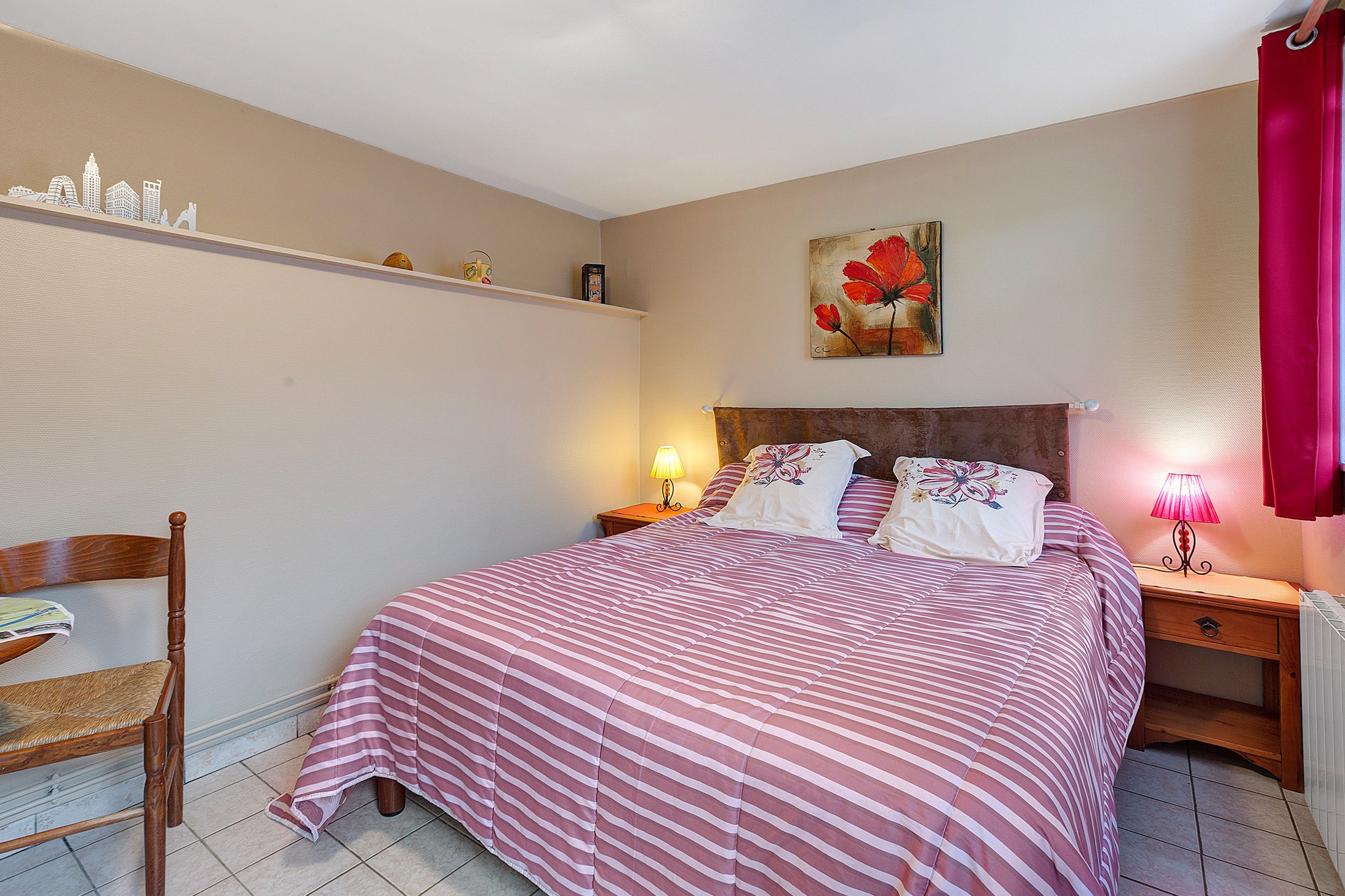 A bedroom with a bed, a chair, a nightstand, a lamp, a shelf with items, and a painting on the wall.