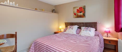 A bedroom with a bed, a chair, a nightstand, a lamp, a shelf with items, and a painting on the wall.