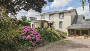 Une maison à deux étages avec une allée en gravier, entourée d’une végétation luxuriante et de plantes en fleurs.