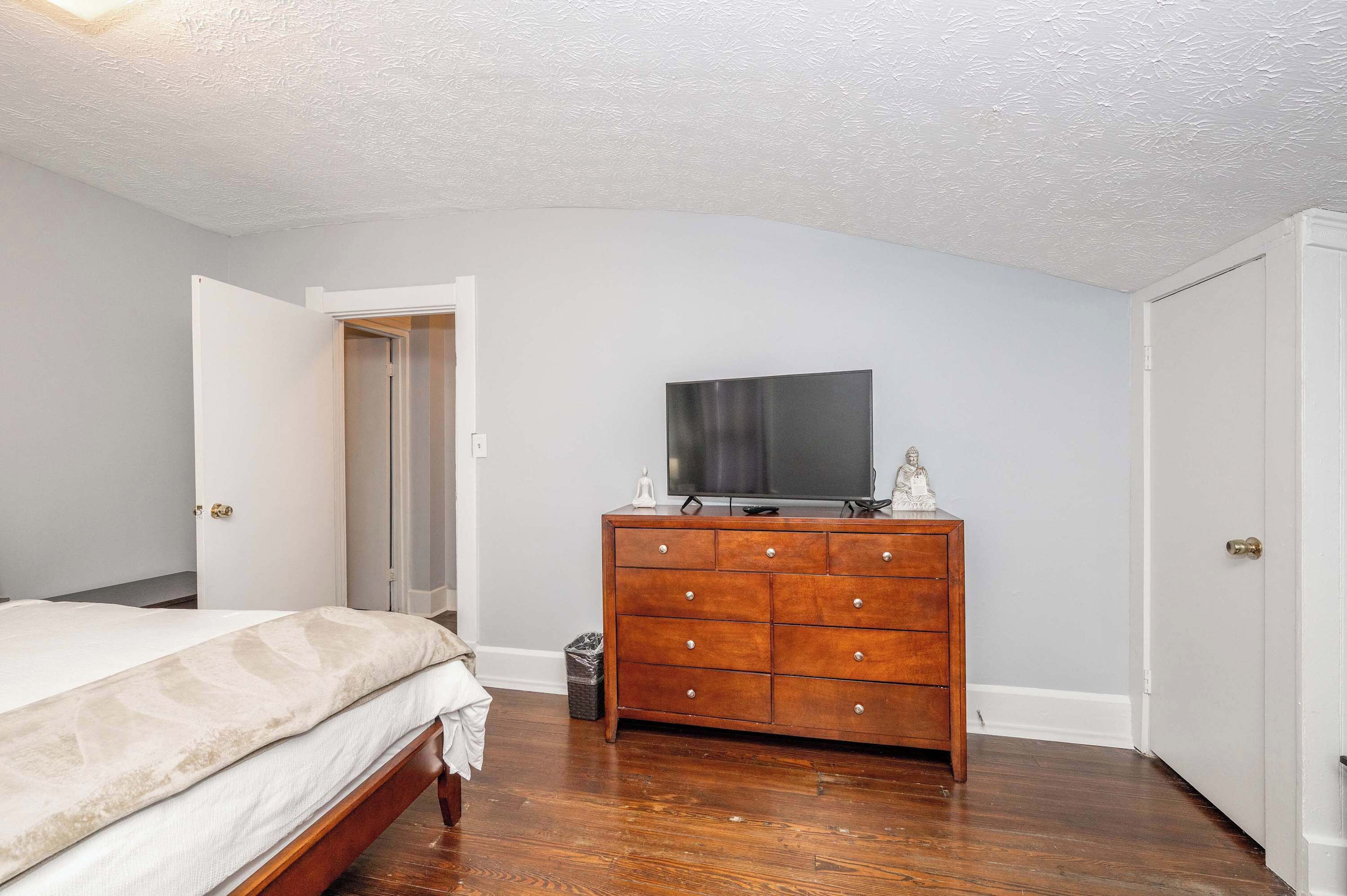 A bedroom with a bed, a wooden dresser, a television on top, and a door leading to another room.