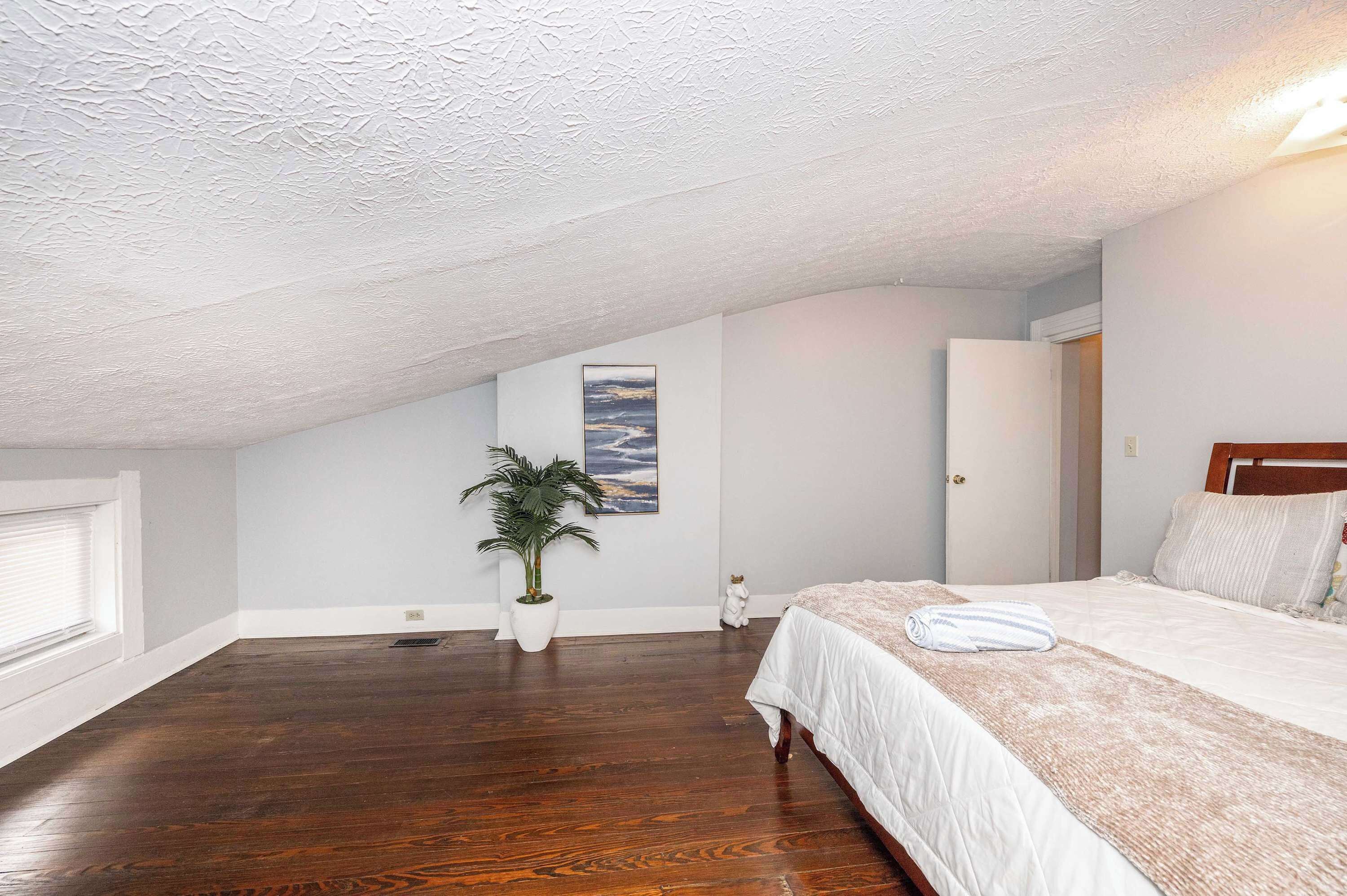 A bedroom with a sloped ceiling, a bed with a white comforter, a potted plant, and a painting on the wall.