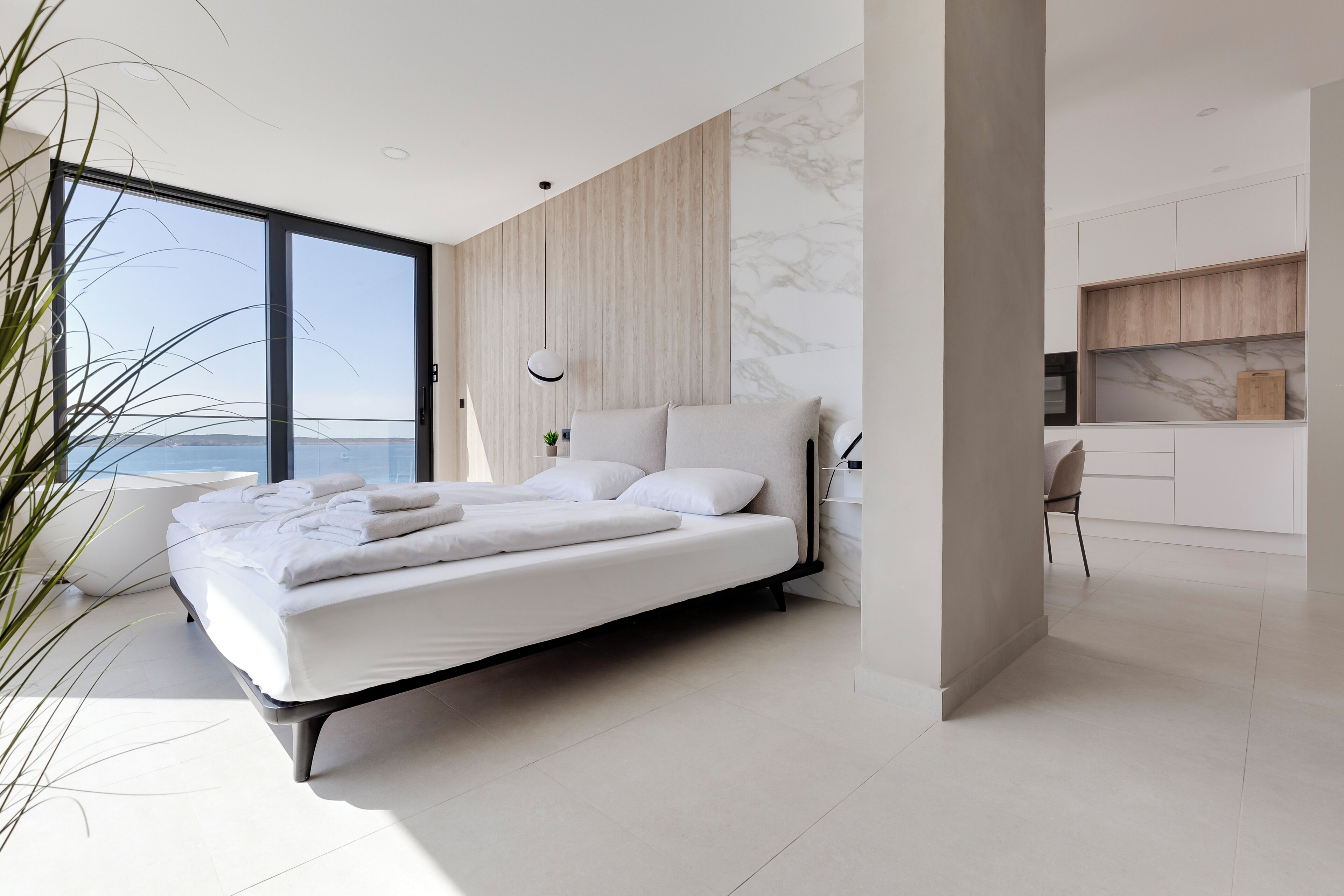Ein modernes Schlafzimmer mit einem großen Bett, Meerblick und einer minimalistischen Küche im Hintergrund.