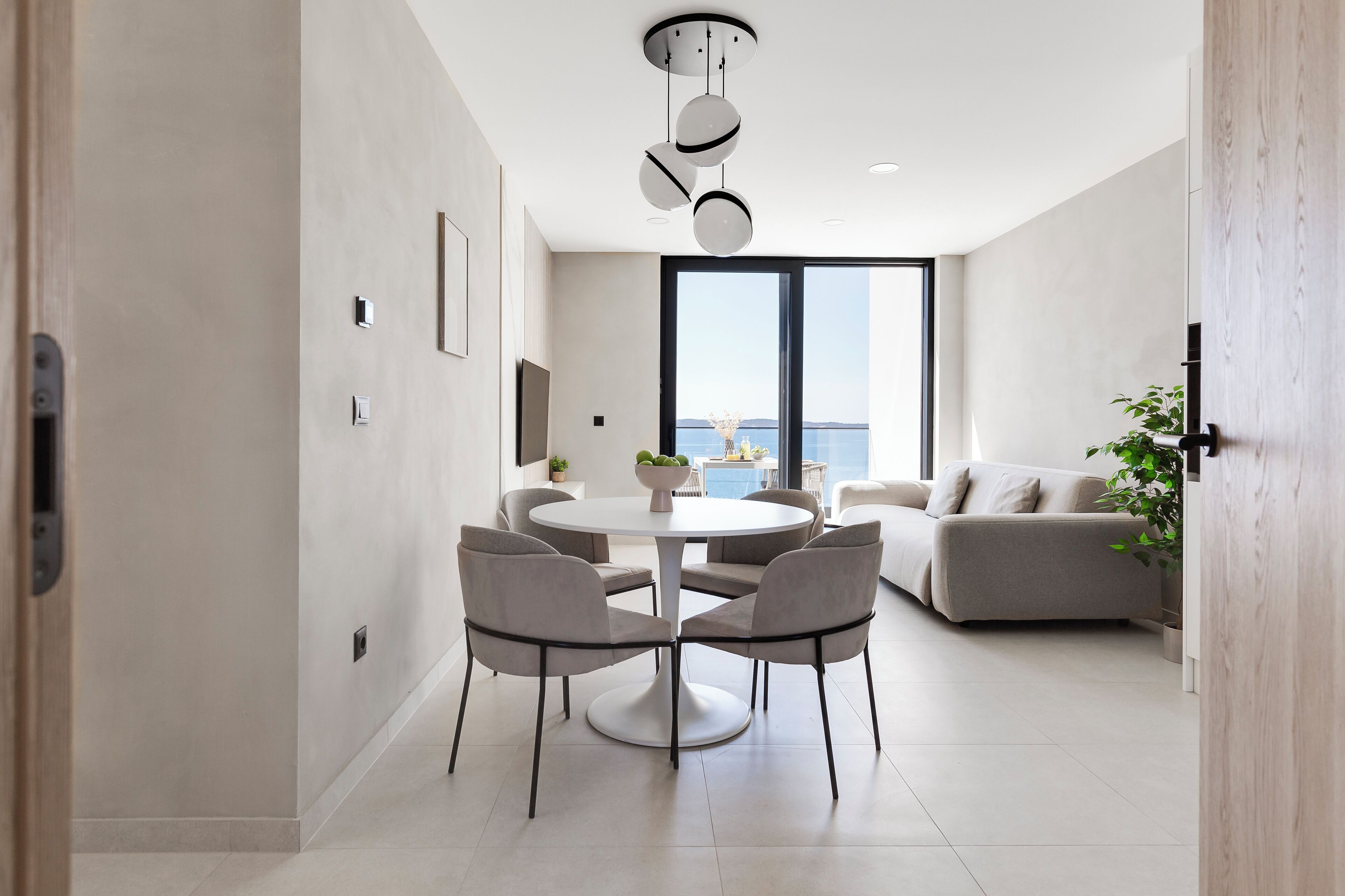 A modern living room with a round dining table, four chairs, a sofa, and a large window offering a view of the sea.