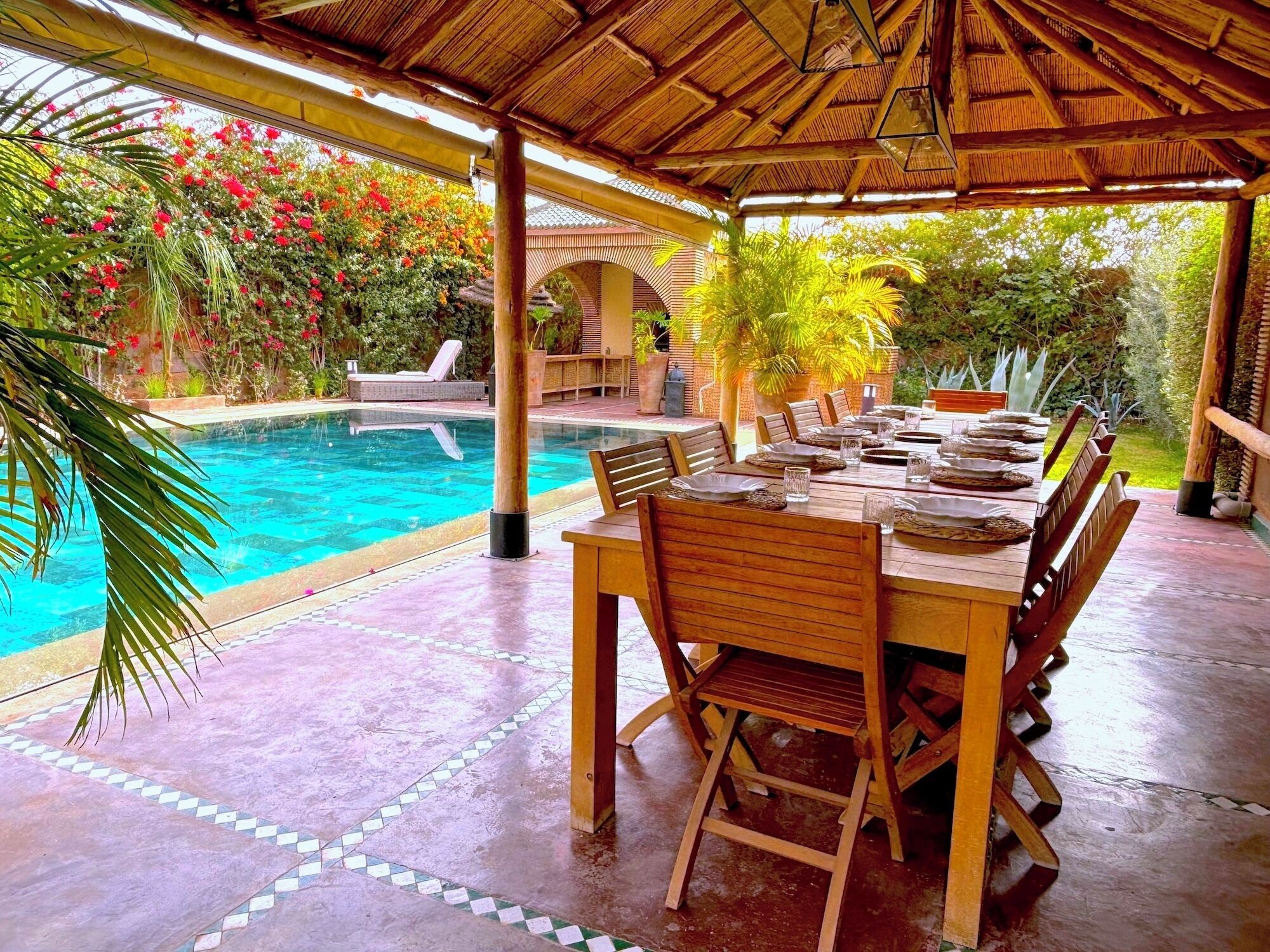 Un espacio de comedor al aire libre cubierto con piscina, mobiliario de madera y abundante vegetación.