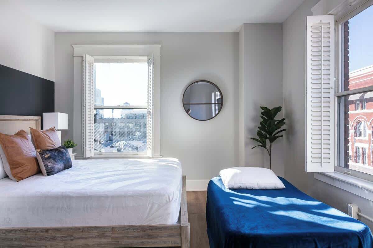 A modern bedroom with a large bed, a smaller bed, a round mirror, and a view of the city through a window.