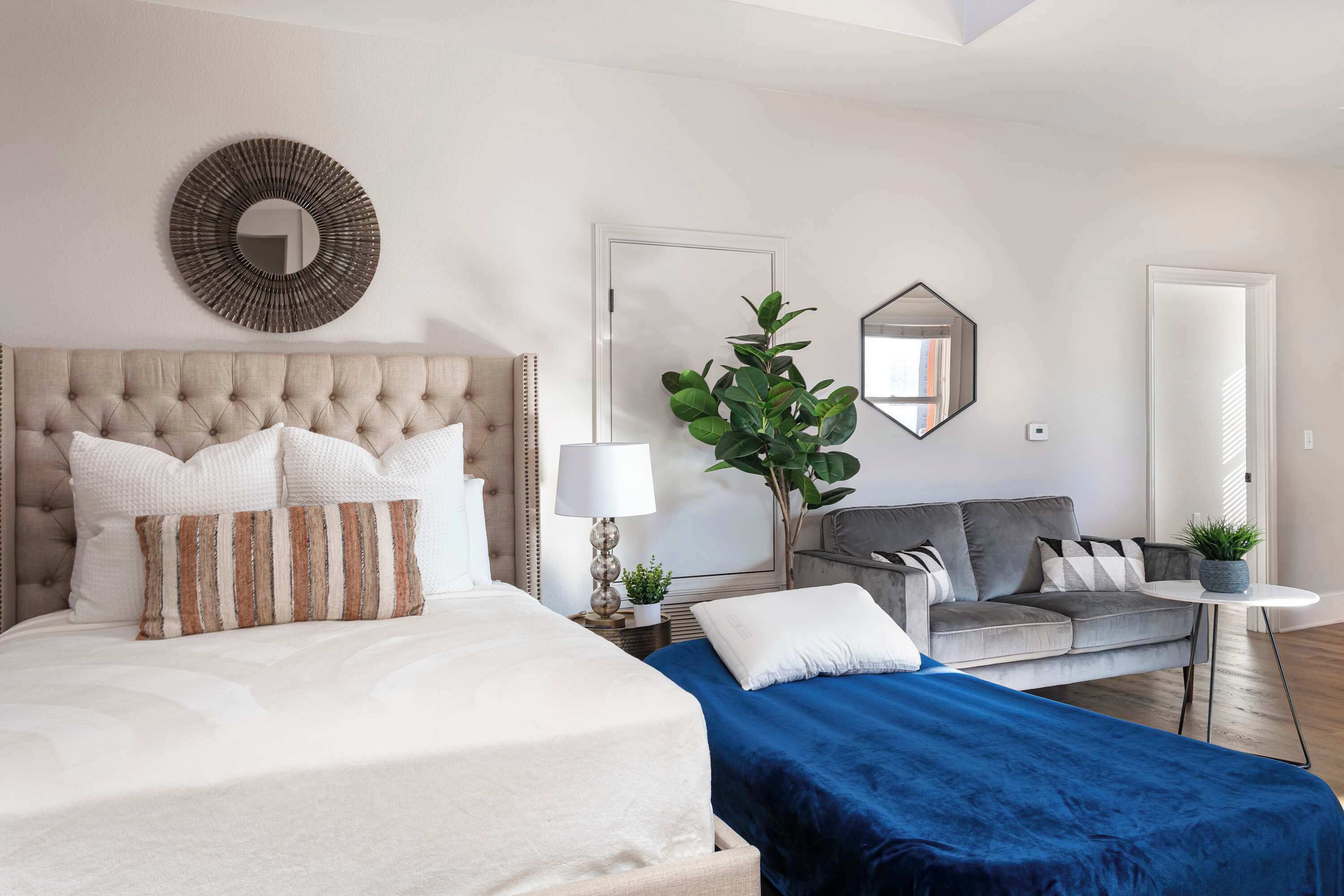 Een slaapkamer met een gebonden hoofdeinde, een bed met witte en gestreepte kussens, een blauwe spreidek, een grijze bank met kussens, een kleine ronde tafel met een plant, en een spiegel aan de muur.