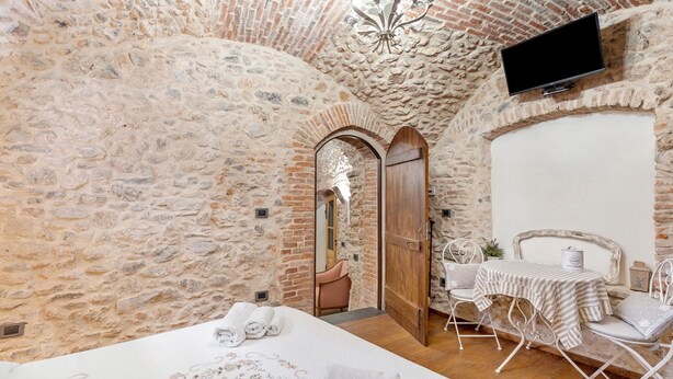 A stone-walled room with a bed, a table with chairs, a TV mounted on the wall, and a chandelier.