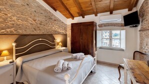 A bedroom with a stone wall, wooden ceiling beams, a bed with towels, a nightstand, a desk, and a television. - Room 'Stanza Della Musica' with Sea View, Shared Terrace and Wi-Fi (CERIALE)