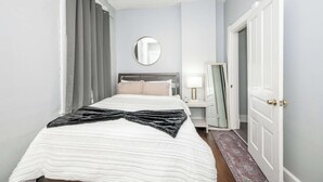 Un lit bien fait, recouvert d’une couverture noire et blanche, une table de chevet avec une lampe, un miroir et une porte sur le côté droit.