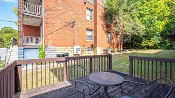 A wooden deck with a table and two chairs, a brick building with a balcony, and a grassy area with trees.