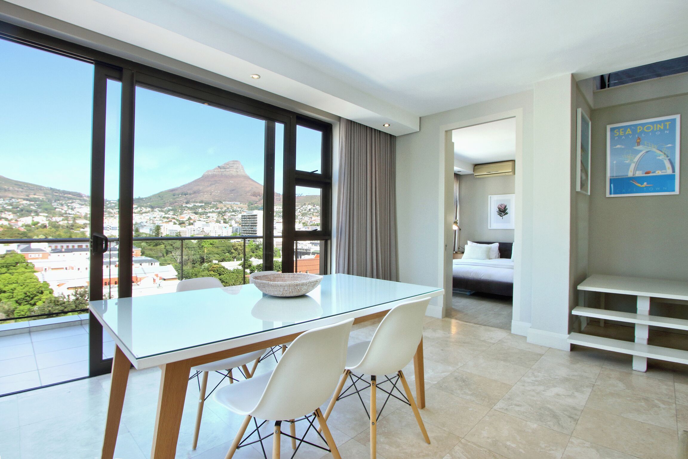 Ein modernes Hotelzimmer mit einem Glastisch, weißen Stühlen, einem Bett und Blick auf Berge und Stadtlandschaft.