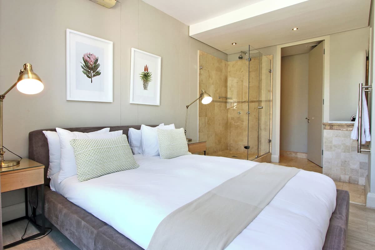 Ein modernes Hotelzimmer mit einem großen Bett, Nachttischlampen, gerahmten Kunstwerken und einer Duschkabine aus Glas.