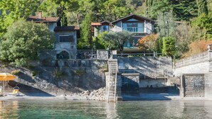 Une propriété située au bord de l’eau, avec un escalier en pierre menant à un pont, une petite piscine et une zone de plage équipée d’une chaise et d’un parasol.