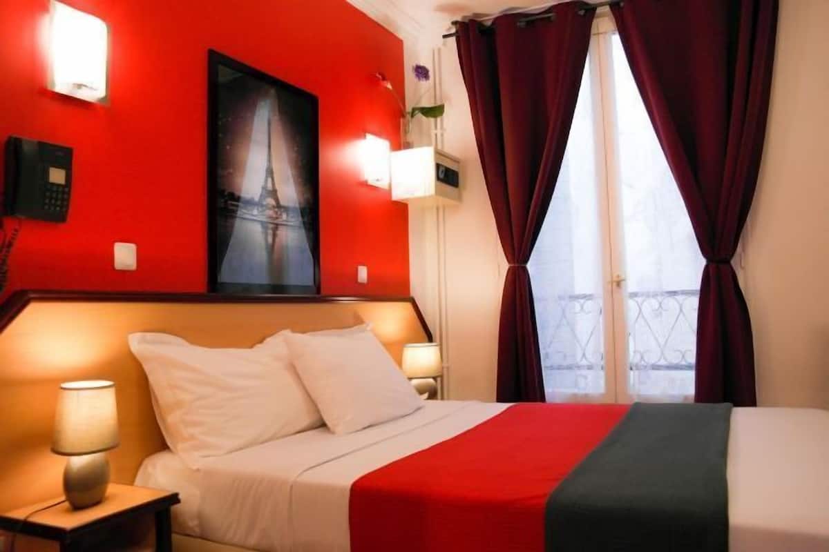 A hotel room with a bed, a nightstand, a lamp, a painting, and red curtains.
