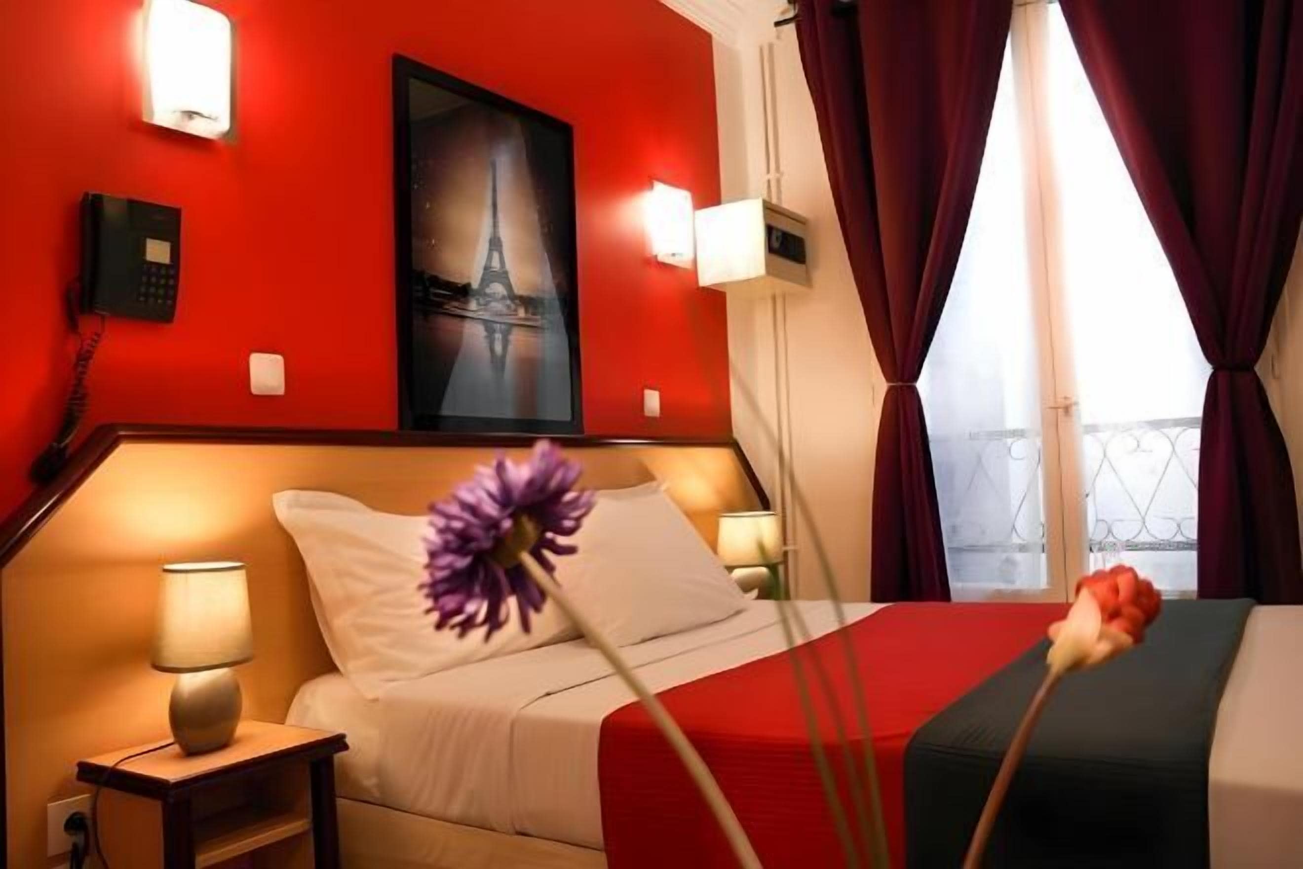 A hotel room with a red wall, a bed with white bedding, a nightstand with a lamp, a painting of the Eiffel Tower, and a window with curtains.