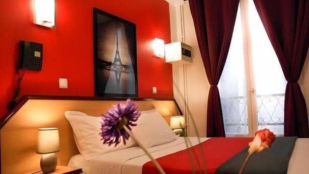 Une chambre d’hôtel avec un mur rouge, un lit avec une literie blanche, une table de chevet avec une lampe, un tableau représentant la Tour Eiffel et une fenêtre avec des rideaux.