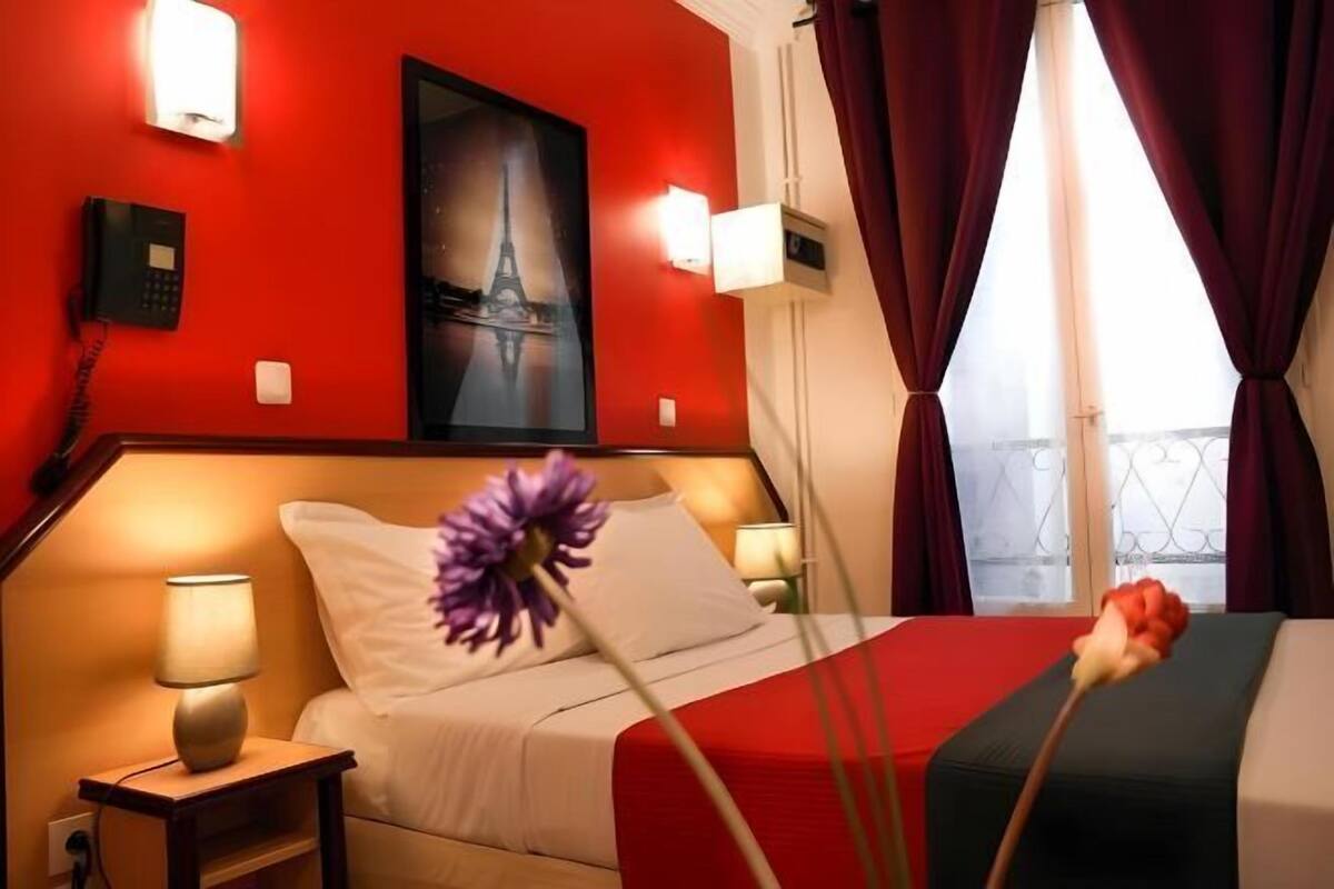A hotel room with a red wall, a bed with white bedding, a nightstand with a lamp, a painting of the Eiffel Tower, and a window with curtains.