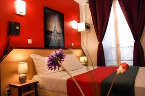 A hotel room with a red wall, a bed with white bedding, a nightstand with a lamp, a painting of the Eiffel Tower, and a window with curtains. - Hotel Audran (Paris)