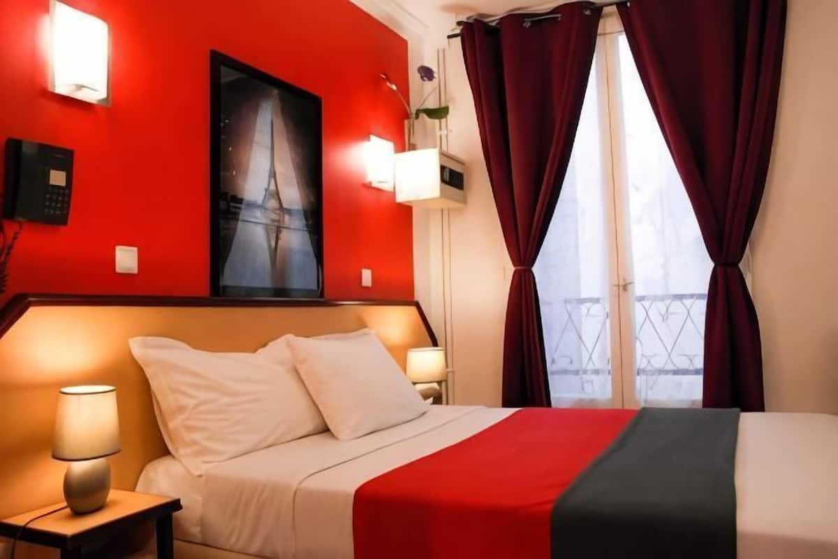 A hotel room with a bed, red walls, a window with curtains, and a painting on the wall.