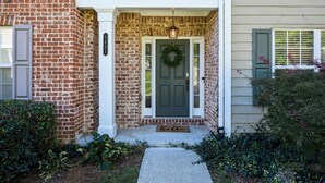 Une maison en briques avec une porte d’entrée vert foncé, un porche blanc et un paillasson sur lequel est écrit « Bonjour ».