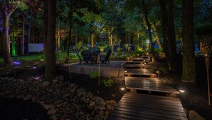 A wooden pathway leading to a seating area with chairs and a table, surrounded by trees and illuminated by soft lighting.