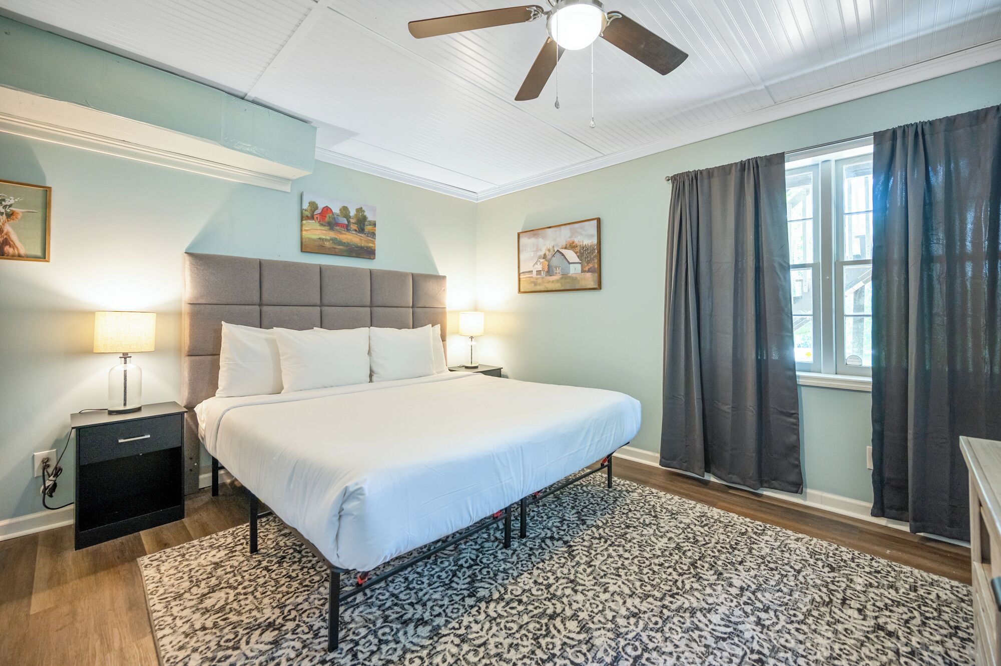 A bedroom with a large bed, two bedside tables with lamps, a ceiling fan, and two paintings on the walls.