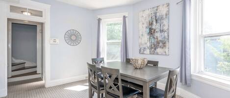 A dining area with a table and chairs, a window with curtains, a wall clock, and a door leading to a staircase.