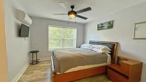 Une chambre à coucher avec un lit, un ventilateur de plafond, une fenêtre, un téléviseur et un petit tabouret.