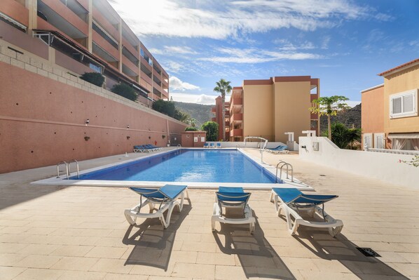 Una piscina con sillones de descanso, un edificio con balcones y un cielo despejado.