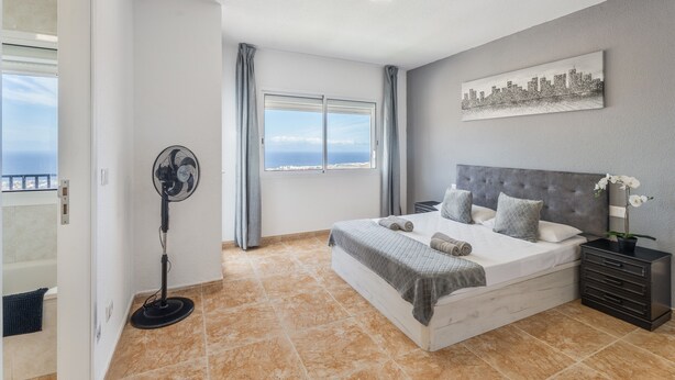 A bedroom with a bed, a fan, a nightstand, a window with curtains, and a cityscape painting on the wall. - Vacation Apartment 'Ocean View Costa Adeje' with Sea View, Private Terrace and Wi-Fi (Costa Adeje)