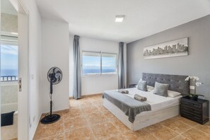 Un dormitorio con una cama, un ventilador, una mesita de noche, una ventana con cortinas y un cuadro de un paisaje urbano en la pared.